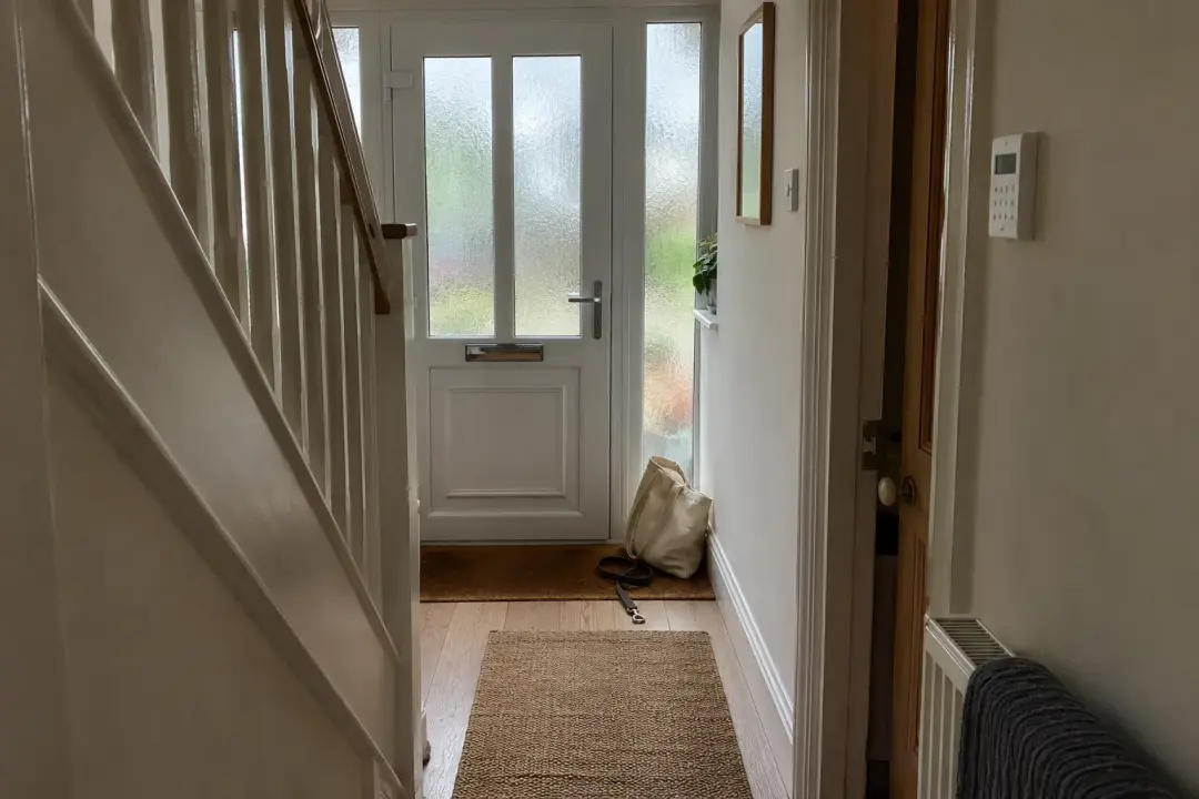 A hallway looking towards a front door, with a shopping bag and a dog lead by the mat and an alarm keypad on the wall